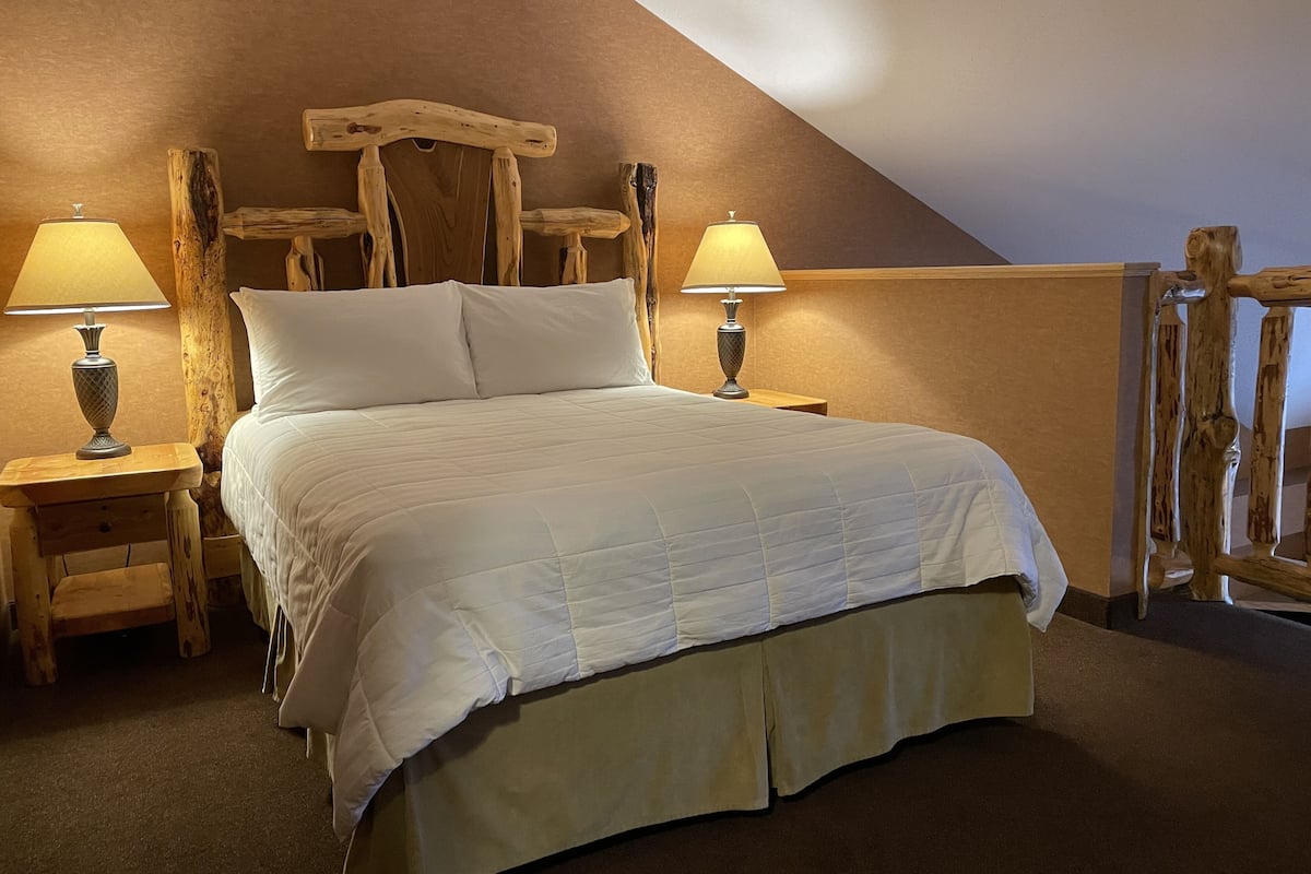 Un dormitorio con cabecera de madera, una cama con ropa de cama blanca, dos mesitas de noche con lámparas y un techo inclinado.