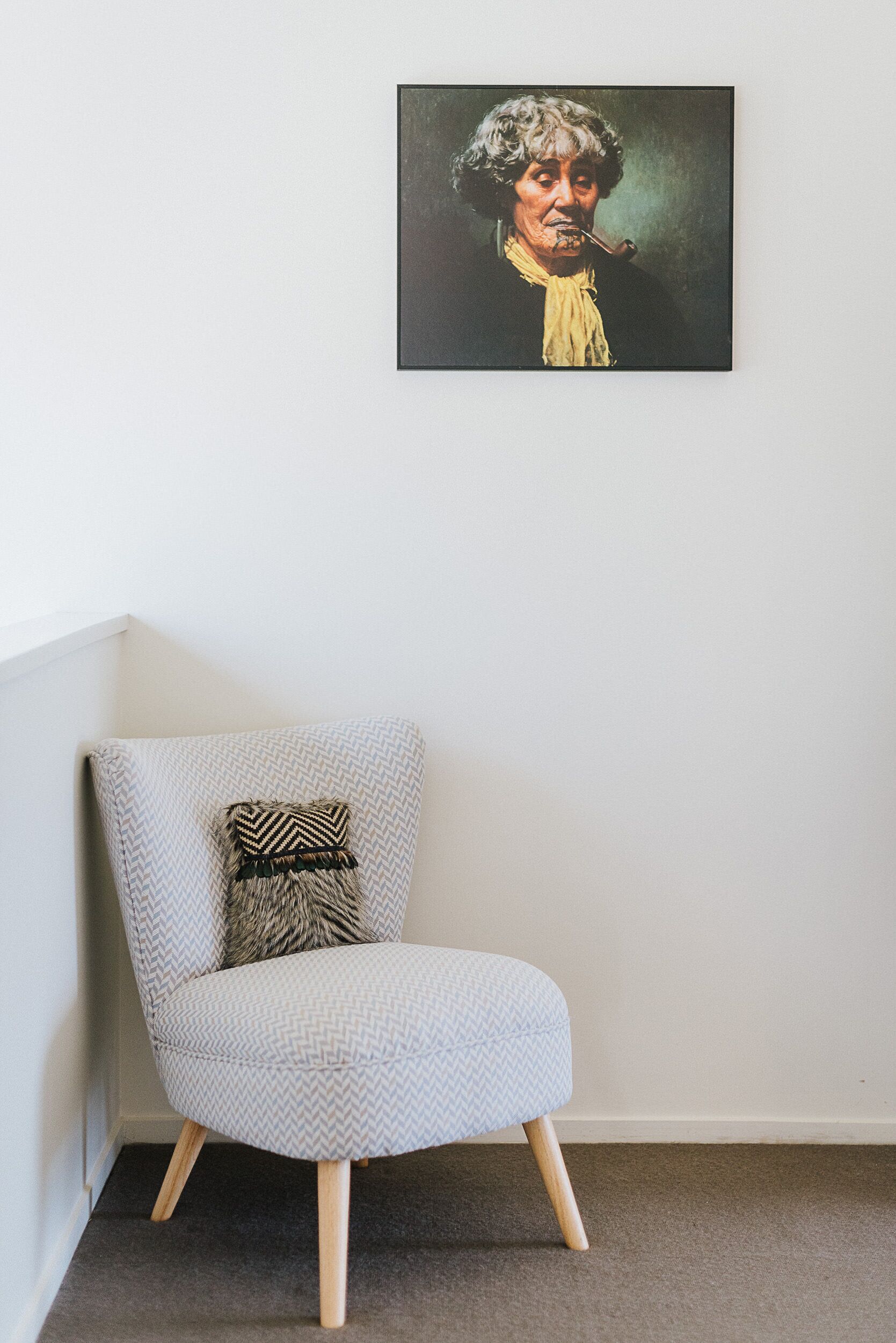 A corner room with a grey patterned armchair, a black and white pillow, and a framed artwork on the wall.