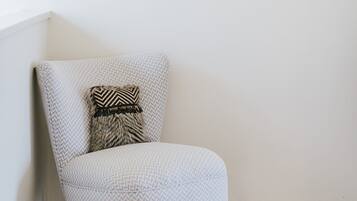 A corner room with a grey patterned armchair, a black and white pillow, and a framed artwork on the wall.