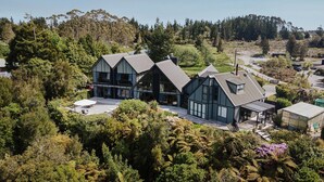A large, modern two-story house with a prominent gabled roof and large windows, surrounded by lush greenery and a hilly landscape.
