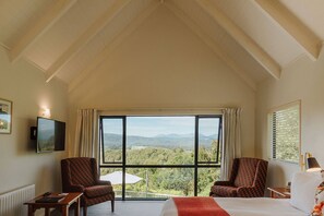 A hotel room with a large window offering a scenic view of the countryside. There are two armchairs and a bed with a red blanket.