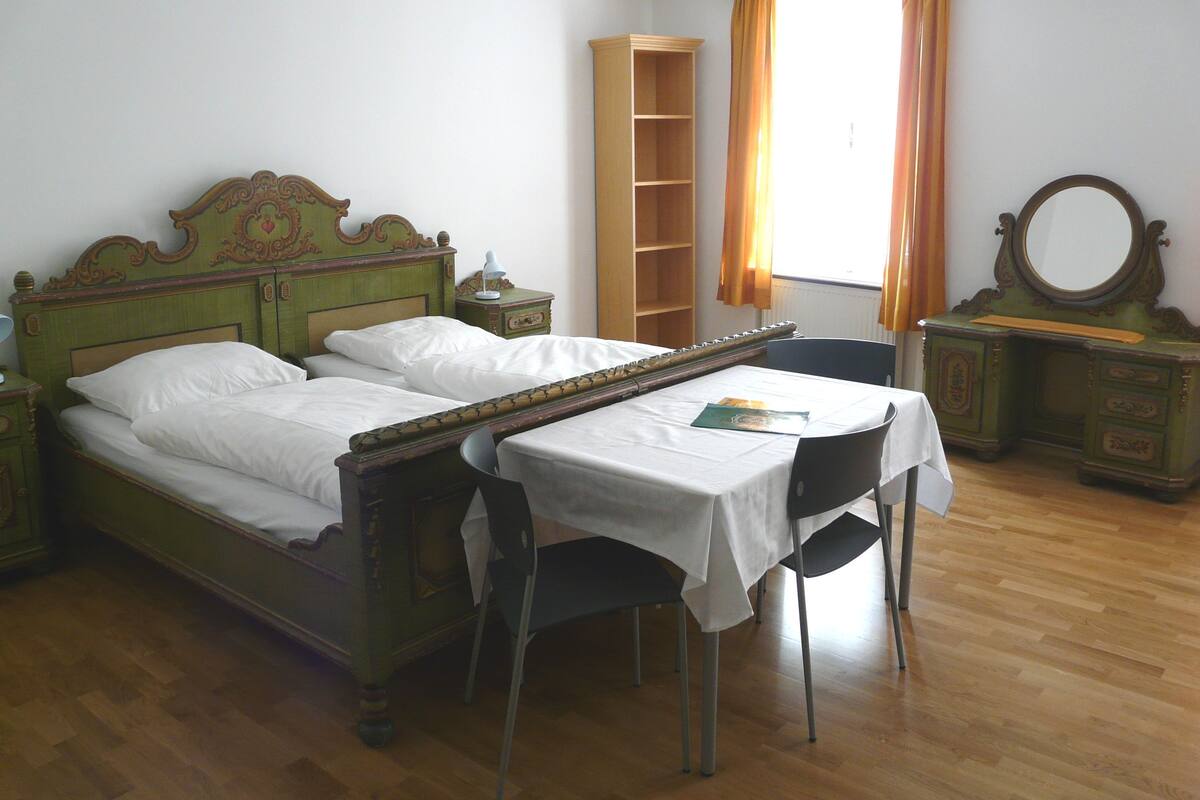 A room with a bed, a table, a chair, a mirror, and a shelf.