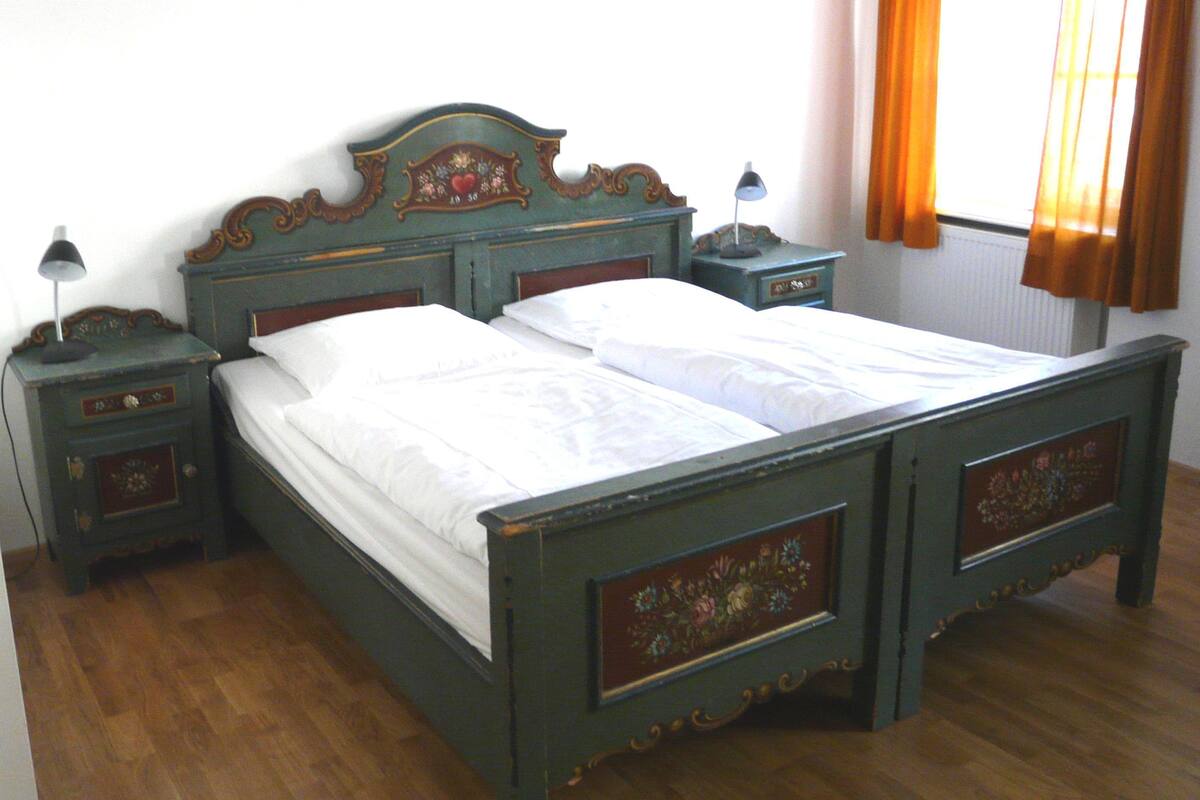 A wooden bed with a floral headboard and footboard, flanked by nightstands with lamps, in a room with wooden flooring and a window with curtains.