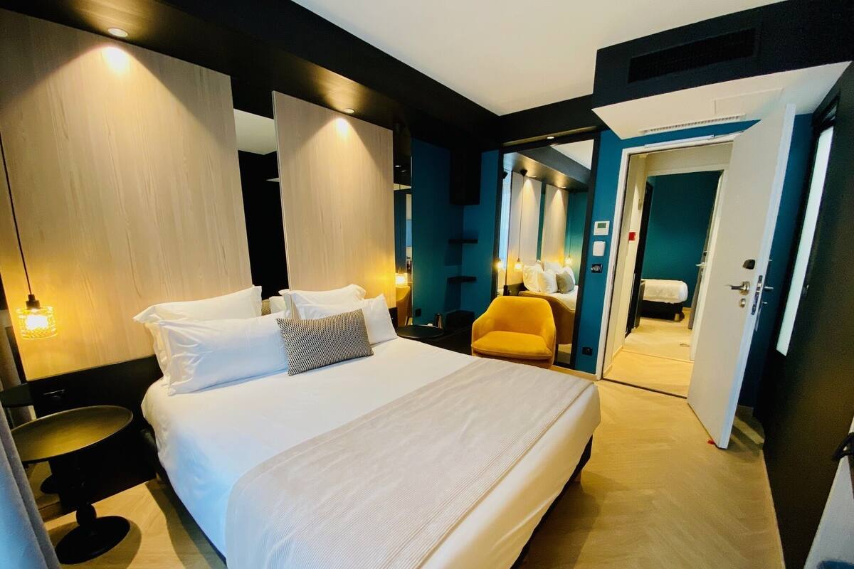 A modern hotel room with a large bed, bedside tables, a chair, and a bathroom visible through an open door.