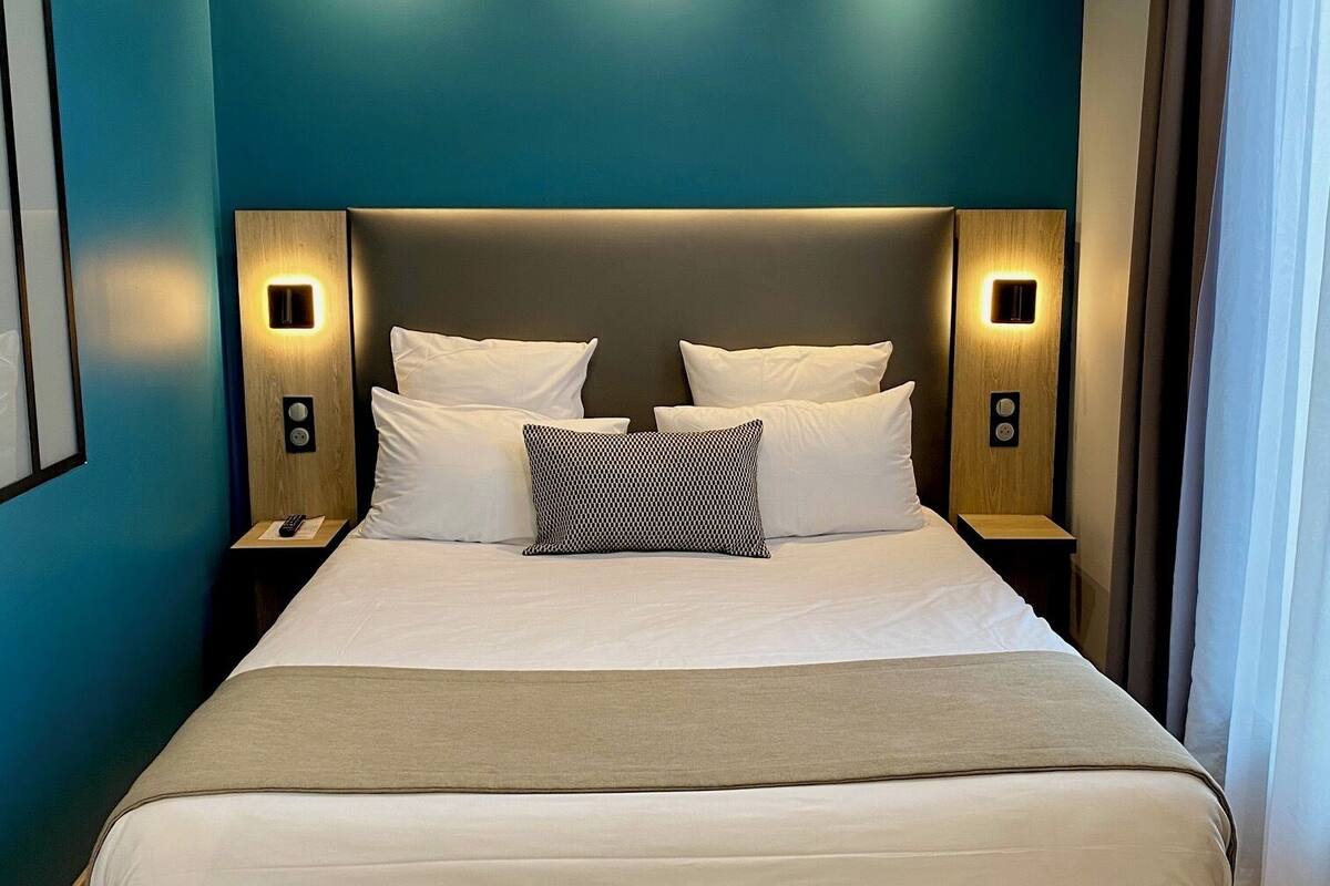 A modern hotel room with a large bed, two bedside tables with lamps, teal walls, and a ceiling light fixture.