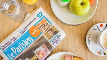 Een ontbijttafel met een krant, een glas sinaasappelsap, een bord met ham, croissants en een fruitschaal met yoghurt en appels.