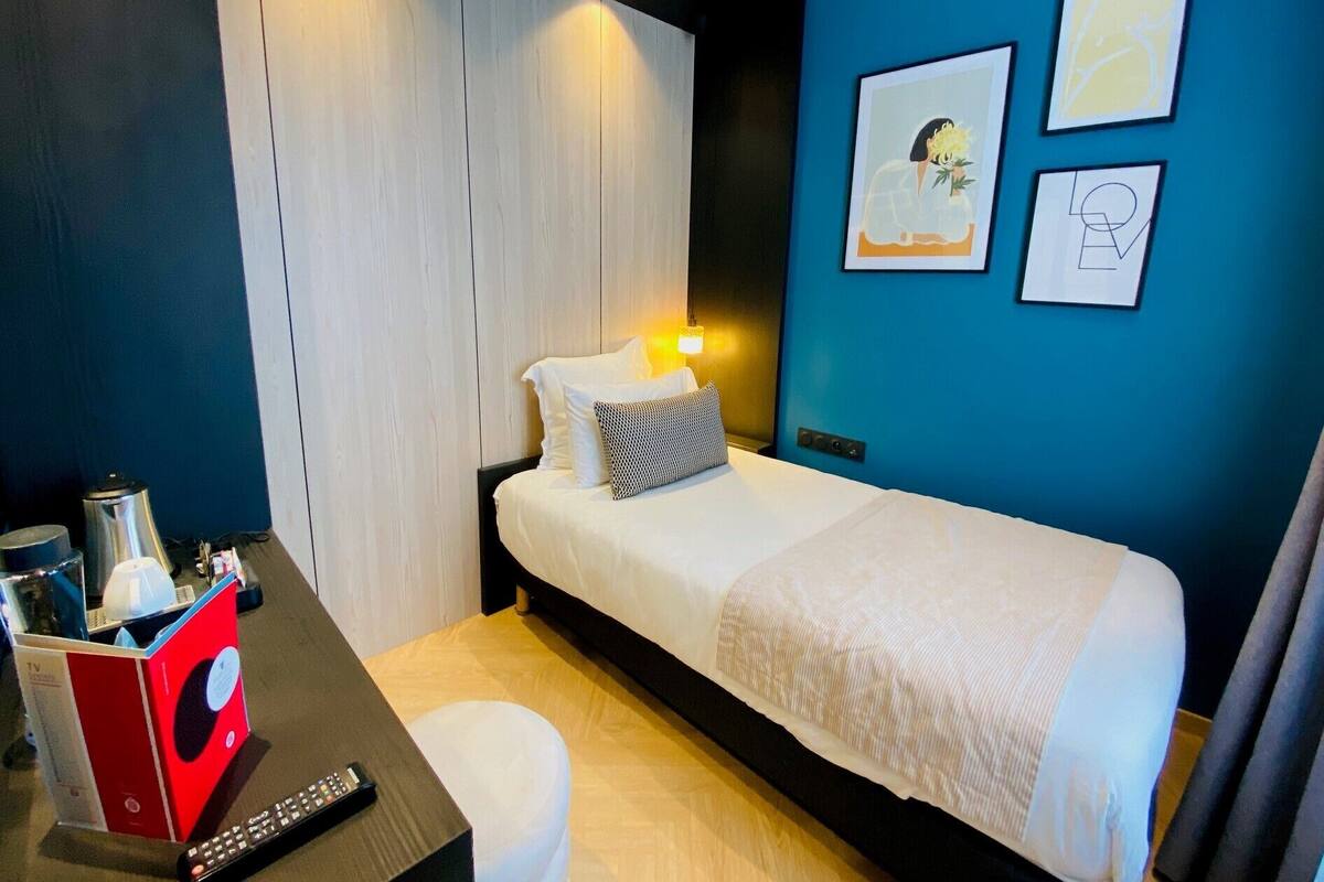 A bedroom with a bed, a bedside table with a remote control, a TV stand, and artwork on the wall.