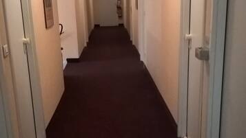 A long, narrow hallway with carpeted floors and white walls. There are doors on both sides, some with signs above them.