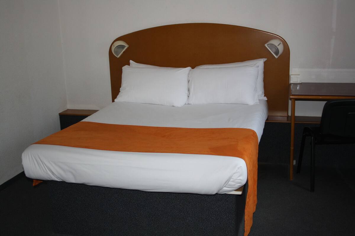 A single bed with white bedding and an orange throw blanket, a wooden headboard, two bedside tables, a desk, and a black chair.