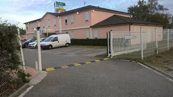 Un hotel con fachada rosa, entrada protegida por un portón y una camioneta blanca estacionada frente a él.