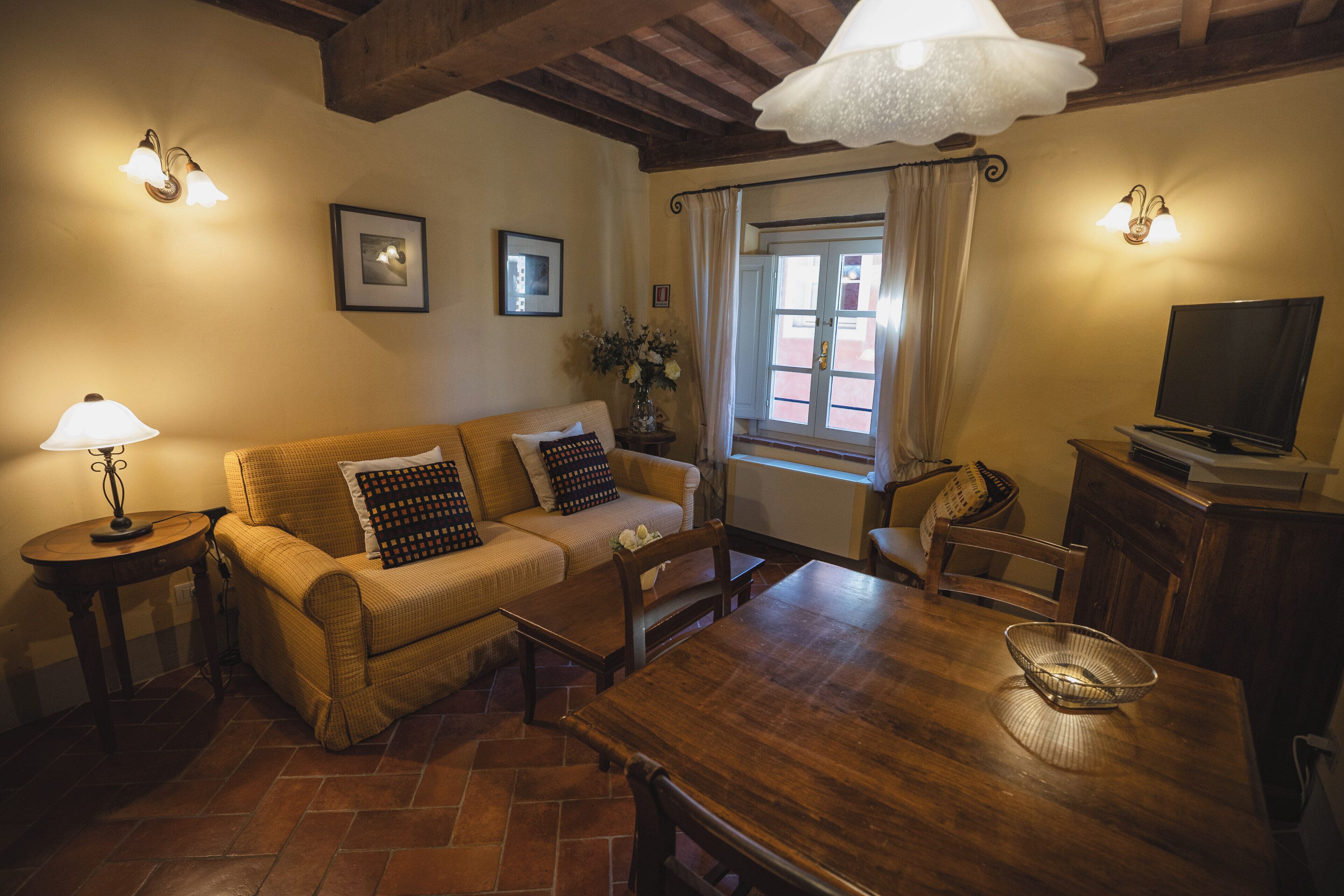 A cozy living room with a wooden dining table, a sofa with patterned cushions, a side table with a lamp, and a television on a wooden cabinet.
