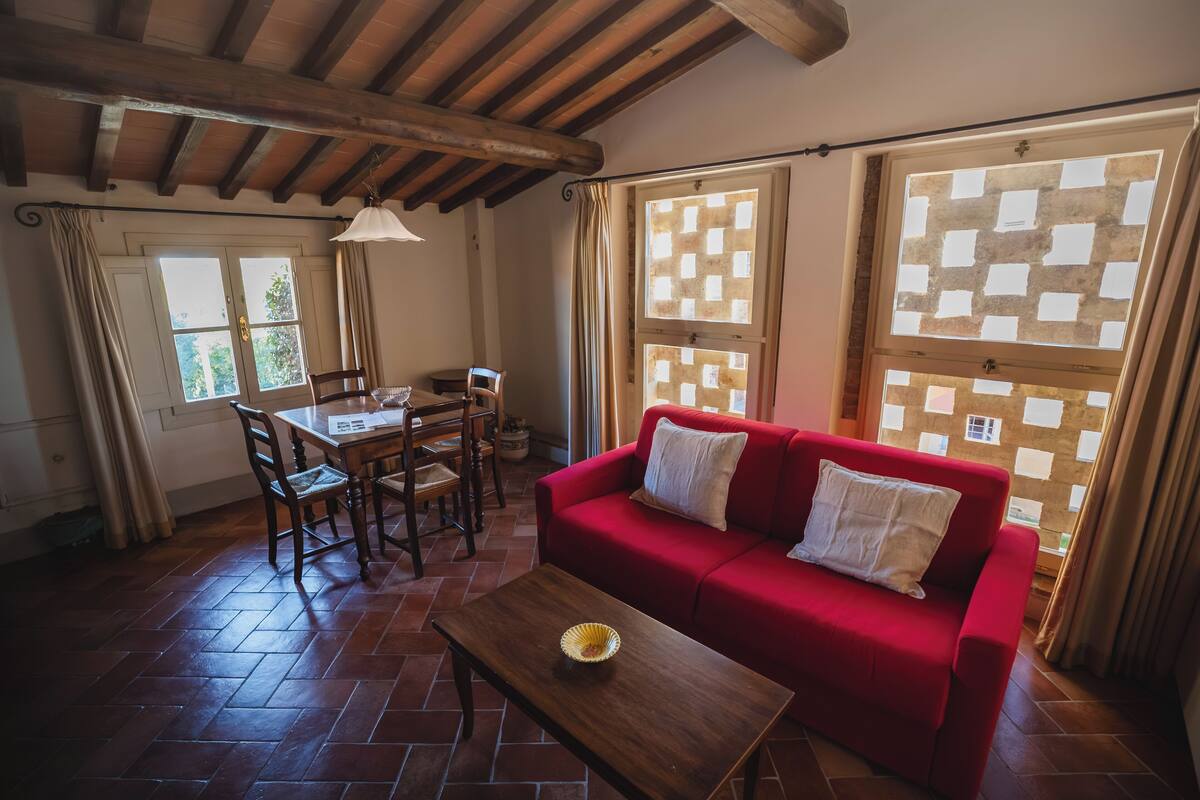 Una sala de estar acogedora con un sofá rojo, una mesa de comedor de madera y sillas. Hay ventanas grandes con cortinas, techo de madera y piso de baldosas.