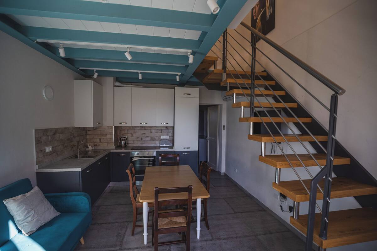 Una cocina compacta con electrodomésticos modernos, un comedor con mesa y sillas de madera, y una escalera con escalones de madera y barandilla de metal.