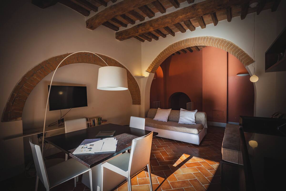 Un área de estar acogedora con una mesa de comedor, sillas, un sofá y un televisor montado en una pared de ladrillo con arco.