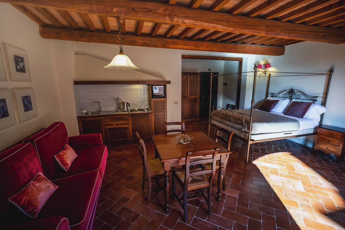 Un cuarto acogedor con techo de madera, un sofá de terciopelo rojo, una mesa de comedor de madera con sillas, una cama con estructura metálica y suelo de baldosas.