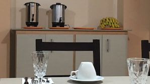 A dining table with a white cup and saucer, a clear glass, and a knife and spoon set. There are two kettles on the counter above and a bunch of bananas on the shelf. - Hotel Mediterráneo Quito (Quito)