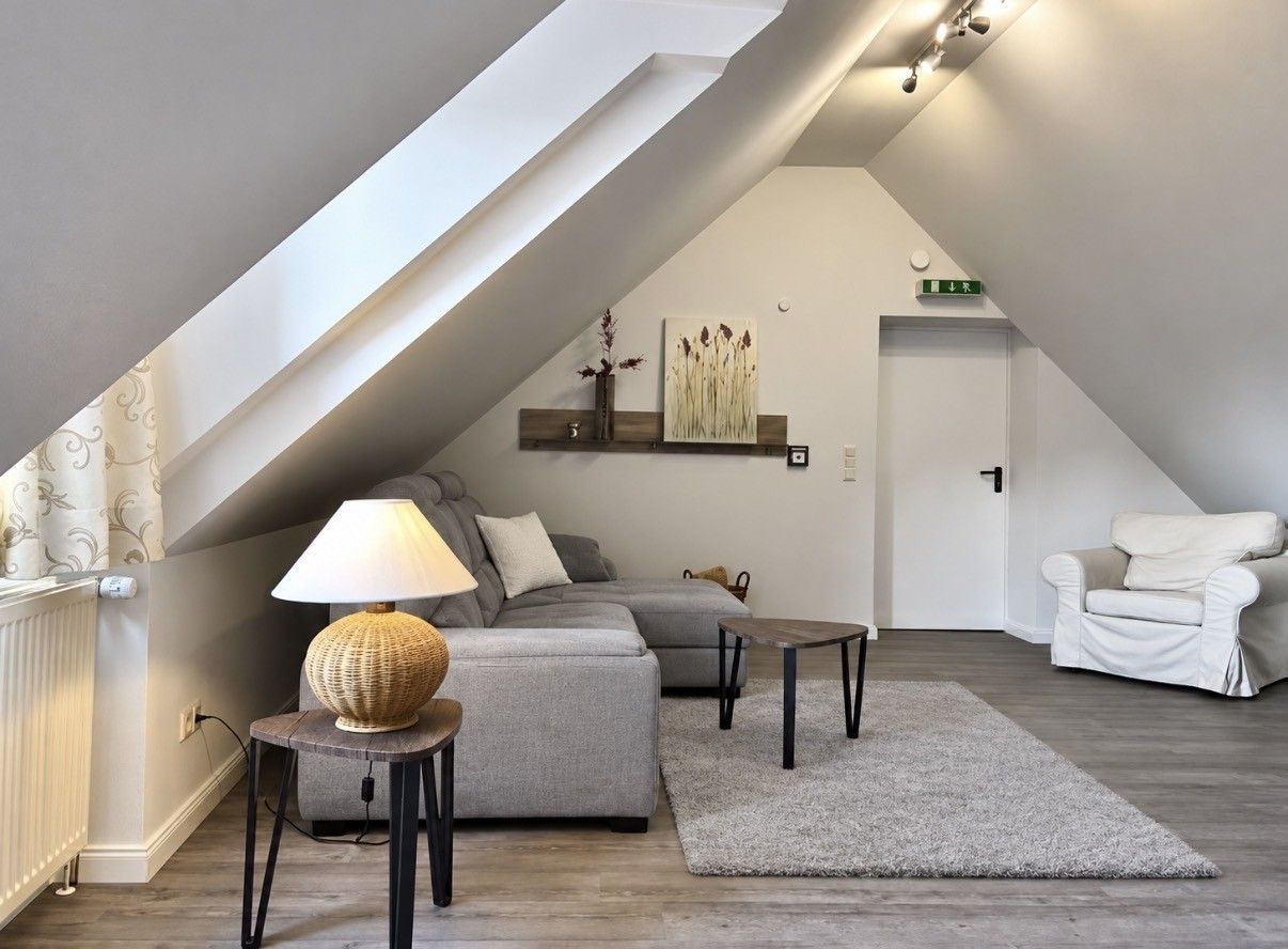 Un dormitorio estilo loft moderno con techo inclinado, un sofá gris, una mesita, una lámpara y una silla blanca.