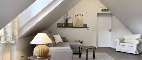 Un dormitorio estilo loft moderno con techo inclinado, un sofá gris, una mesita, una lámpara y una silla blanca.