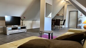 Ein modernes Wohnzimmer mit Flachbildfernseher, einer Couch mit Kissen, einem Essbereich mit Stühlen und einer Küche im Hintergrund.