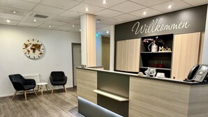 Ein Empfangsbereich mit einem Holzbüro, zwei schwarzen Stühlen, einer Wanduhr und einem Schild mit der Aufschrift 'Willkommen'.
