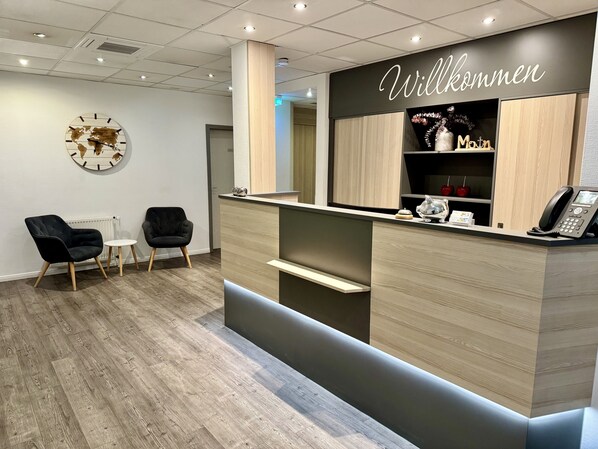 Un espace d’accueil avec un comptoir en bois, deux chaises noires, une horloge au mur et un panneau indiquant « Willkommen ».