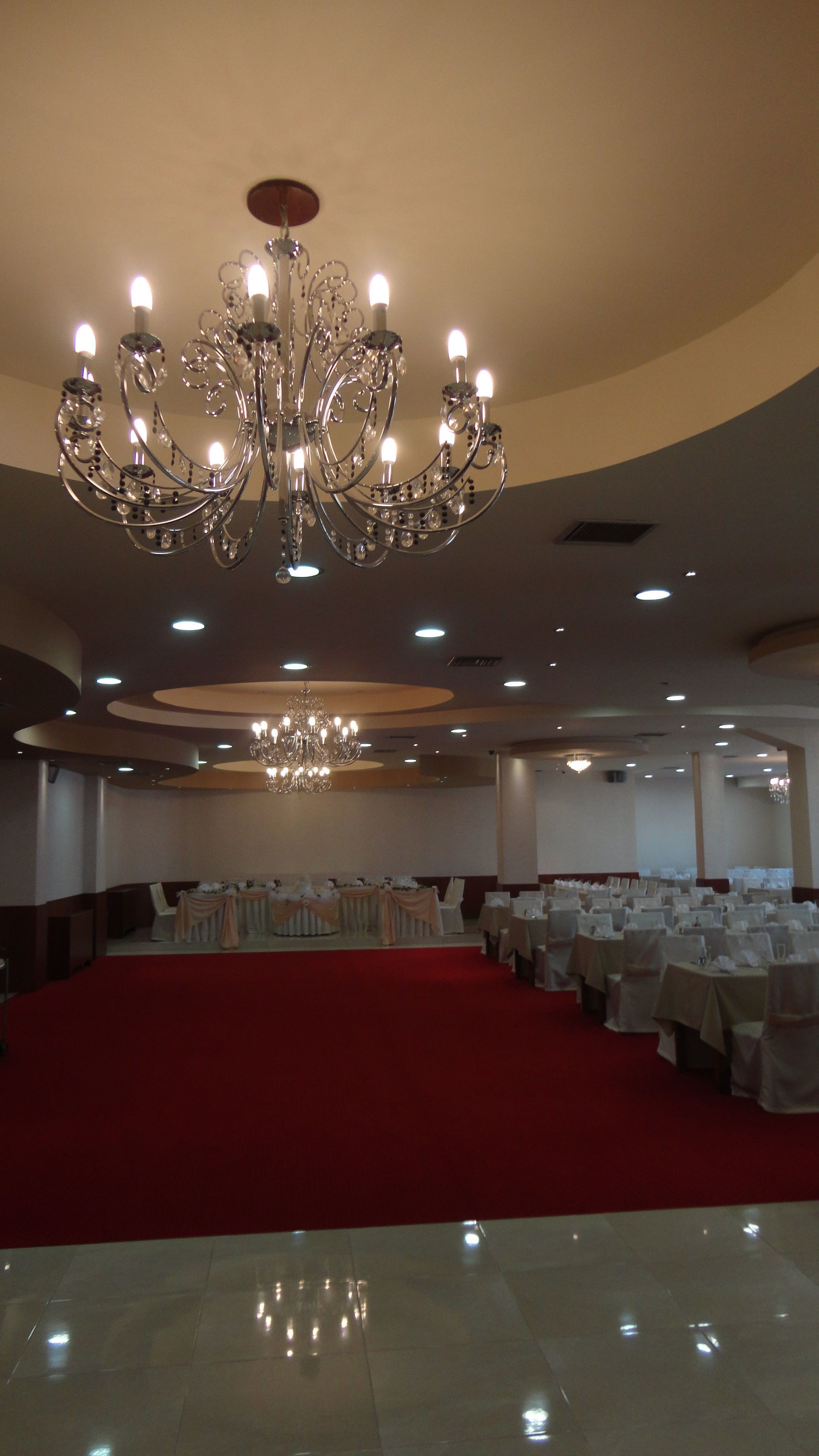 Un'ampia sala con un grande lampadario, diversi tavoli con tovaglie bianche e un tappeto rosso.