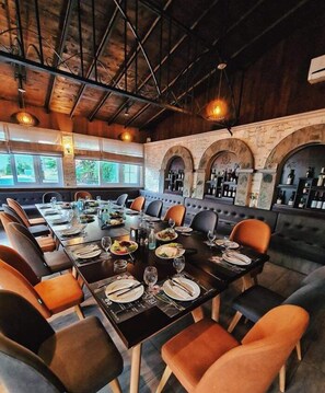 Un restaurante con una larga mesa común, asientos acolchados y una zona de bar con estanterías llenas de botellas.
