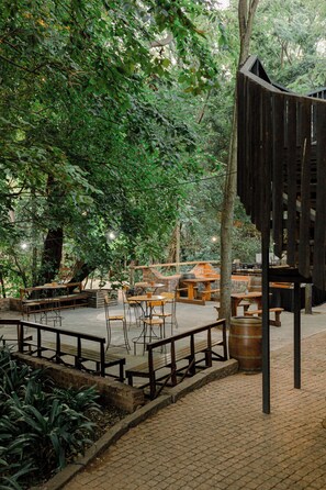 An outdoor seating area with tables and benches, surrounded by trees and a brick wall. - Kleinkaap Boutique Hotel (Centurion)