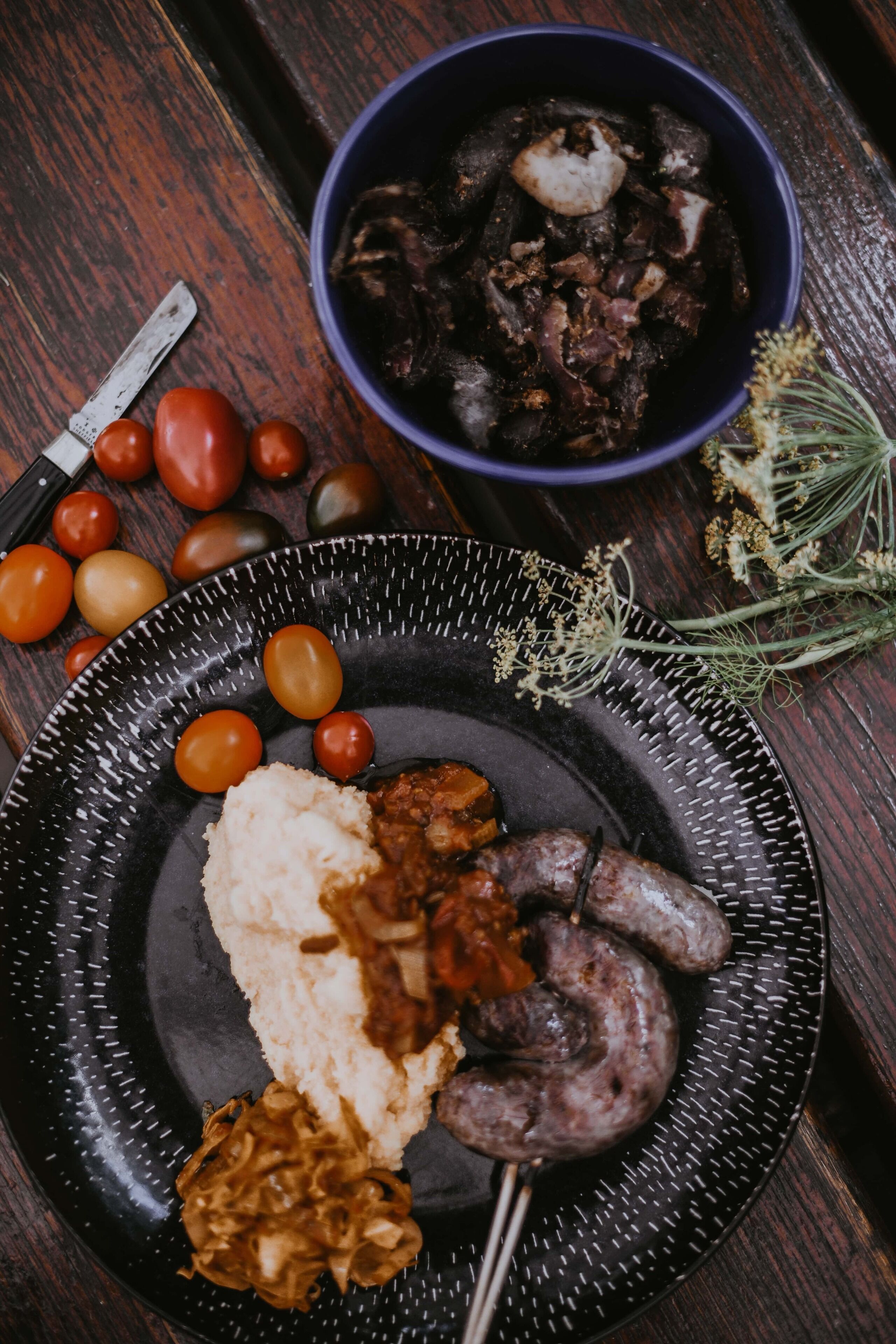 A plate with sausages, mashed potatoes, and a side of pickled vegetables on a dark speckled plate.