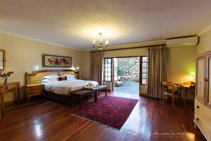 A bedroom with a wooden floor, a bed, a nightstand, a desk, and a chair. There is a window with curtains, a chandelier, and a painting on the wall. - Kleinkaap Boutique Hotel (Centurion)