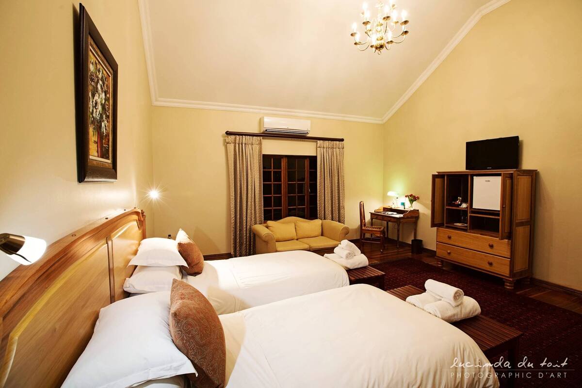 A hotel room with two beds, a wooden headboard, a sofa, a TV, a desk, and a chandelier.