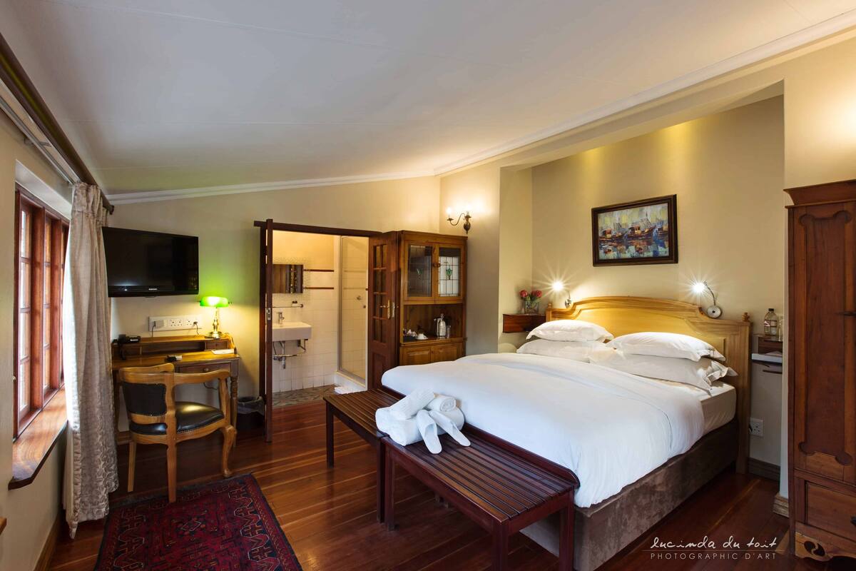 A hotel room with a bed, a desk, a chair, a bench, a TV, a painting, and a wardrobe.