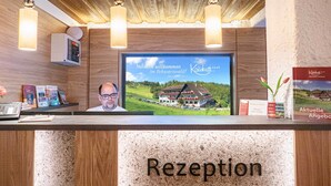A reception desk with a wooden finish, a mounted television displaying a welcome message, and a framed photo of a building. - Höhenhotel & Restaurant Kalikutt (Oppenau)