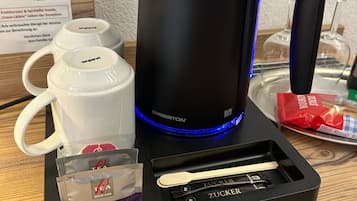 A coffee maker with a black base and a stainless steel pot, accompanied by white mugs and tea packets.