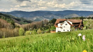 A house with a sloped roof, surrounded by lush green fields and mountains in the background. - Höhenhotel & Restaurant Kalikutt (Oppenau)