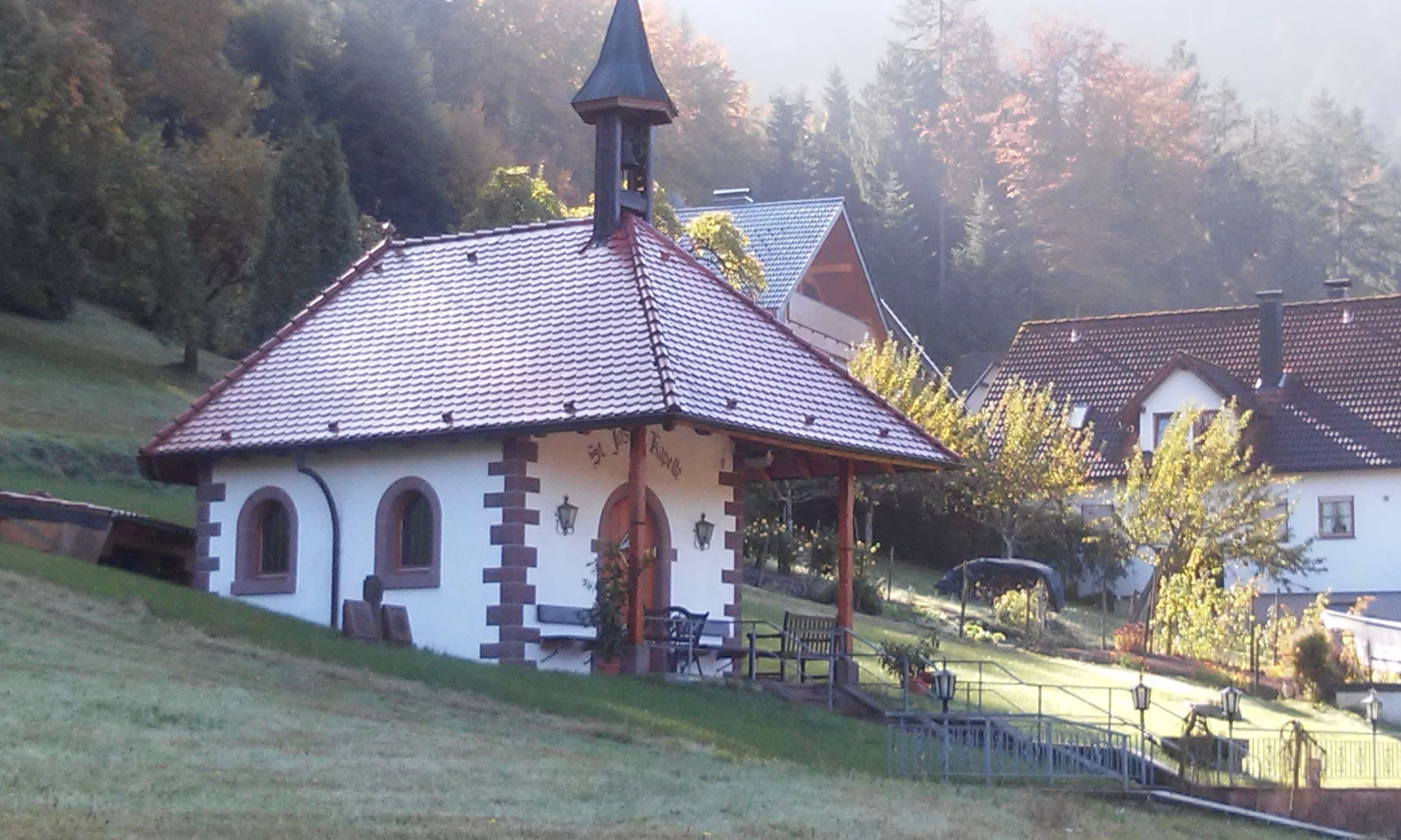 A small chapel with a tiled roof and a bell tower, surrounded by a grassy area and trees.