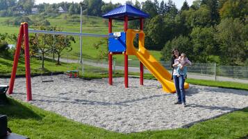 A playground with a slide and swings, surrounded by a fence and greenery.