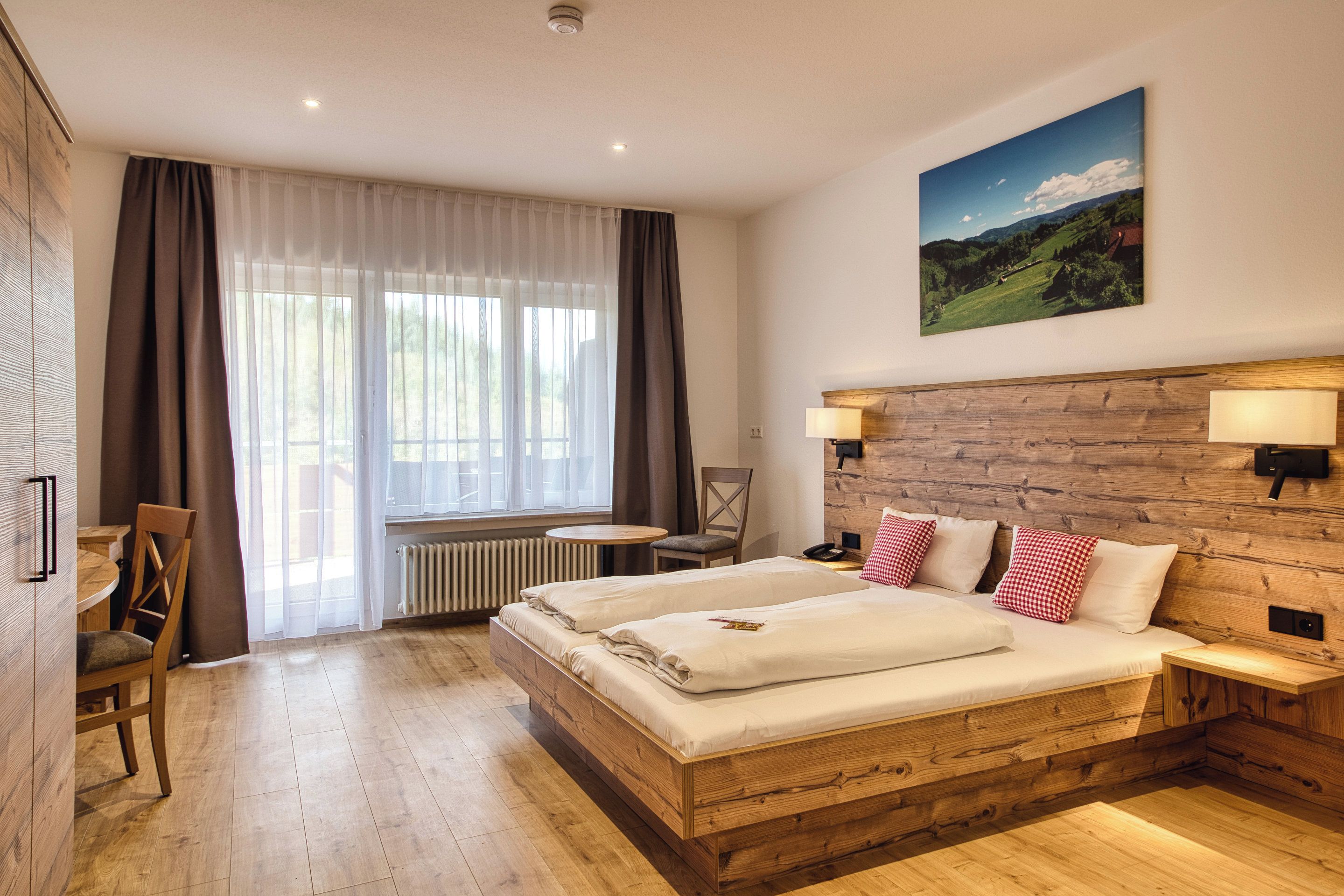 Une chambre d’hôtel avec un lit en bois, un bureau, une chaise, un radiateur et un tableau accroché au mur.