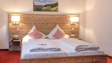 A double bed with white linens and red and white checkered pillows, a wooden headboard, two bedside lamps, a nightstand with a phone, and a painting of a landscape on the wall.
