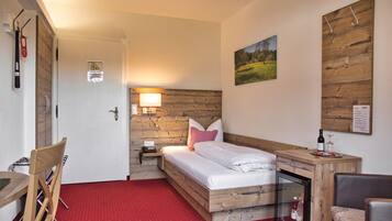 Ein kompaktes Hotelzimmer mit einem hölzernen Kopfteil, einem Einzelbett mit weißen Laken, einem kleinen Nachttisch mit Lampe, einem Teppich mit roten Punkten und einer Holzvertäfelung.