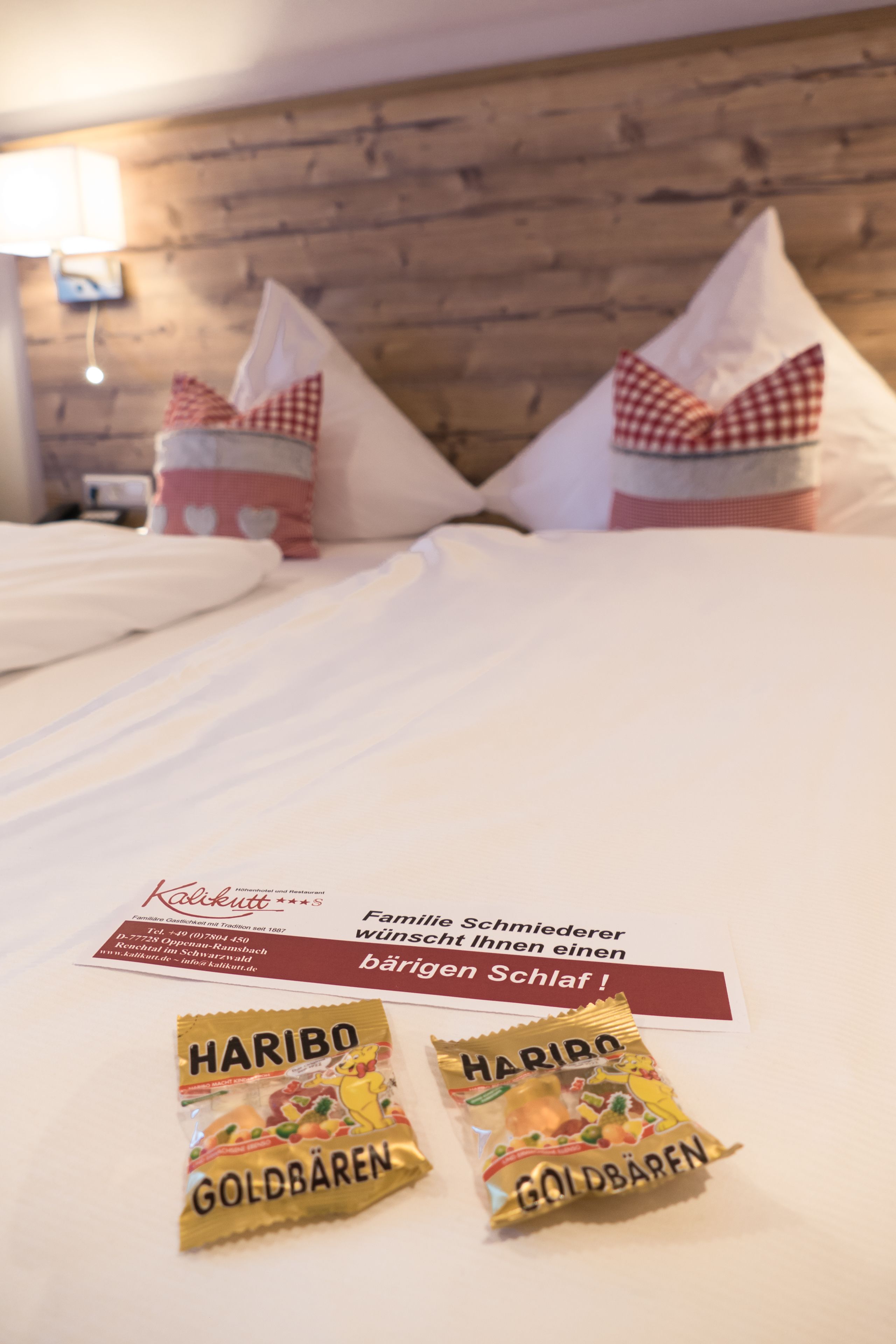 Et hotelværelse med sengegavl i træ, en seng med hvide sengetøj og to puder med røde og hvide tern. Der ligger to pakker Haribo Goldbären på sengen.