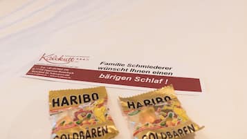 Quarto de hotel com cabeceira de madeira, cama com roupa de cama branca e duas almofadas com padrão xadrez vermelho e branco. Há dois pacotes de Haribo Goldbären na cama.
