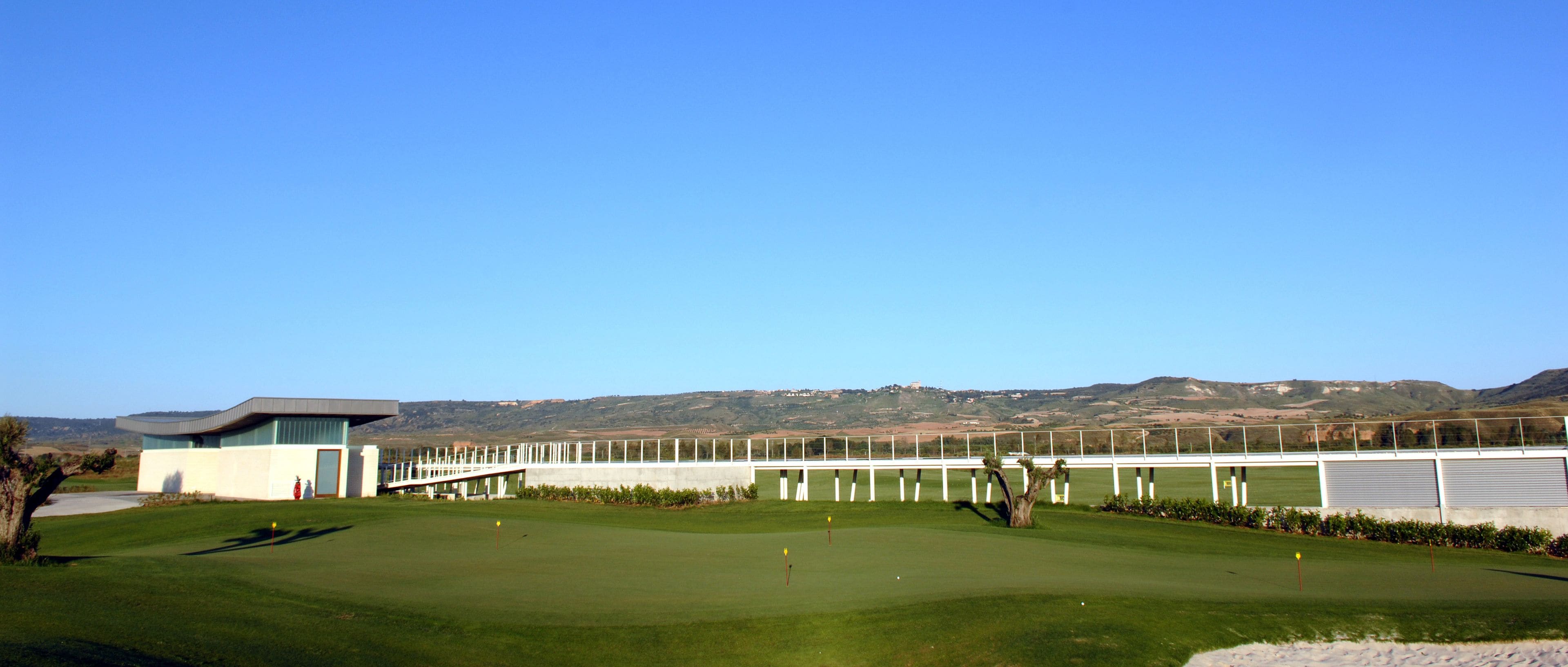 Un campo de golf con un moderno club de campo, un puente y un telón de fondo de montañas panorámicas.