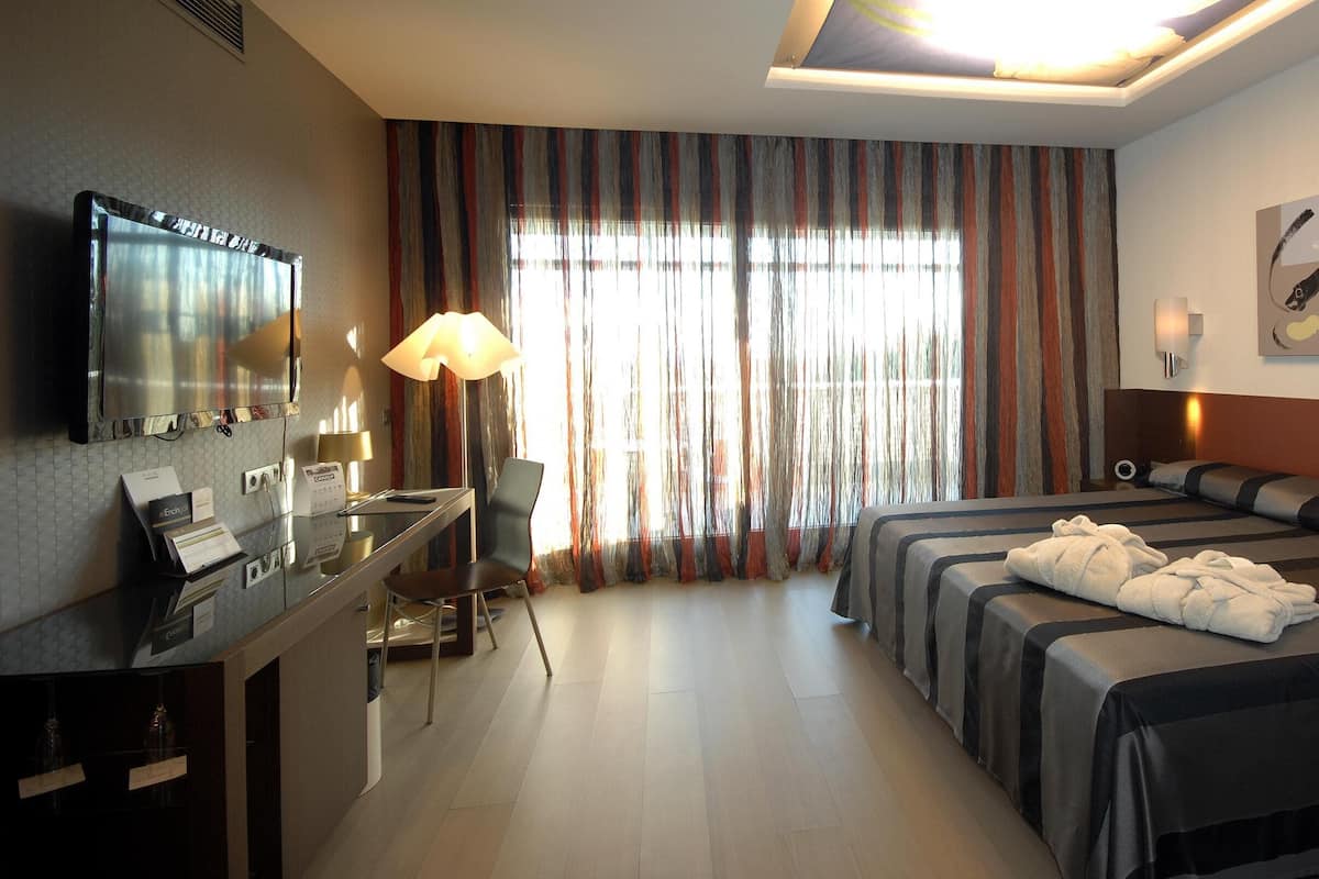 A hotel room with a bed, a desk with a lamp, a television, and a large window with curtains.