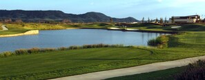 A golf course with a pond, a walking path, and a modern building in the background. - Sercotel El Encin Golf (Alcala de Henares)