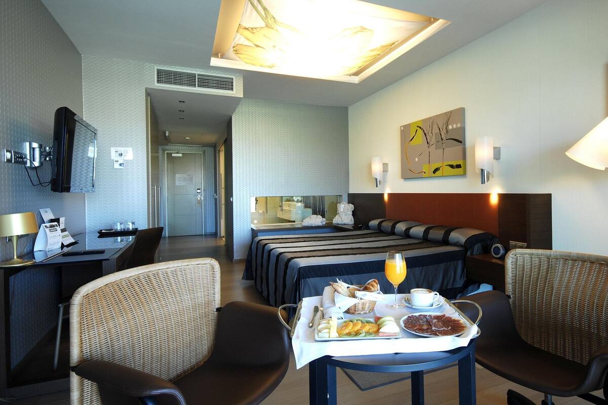 A hotel room with a bed, two chairs, a table with breakfast, a TV, and artwork on the wall.
