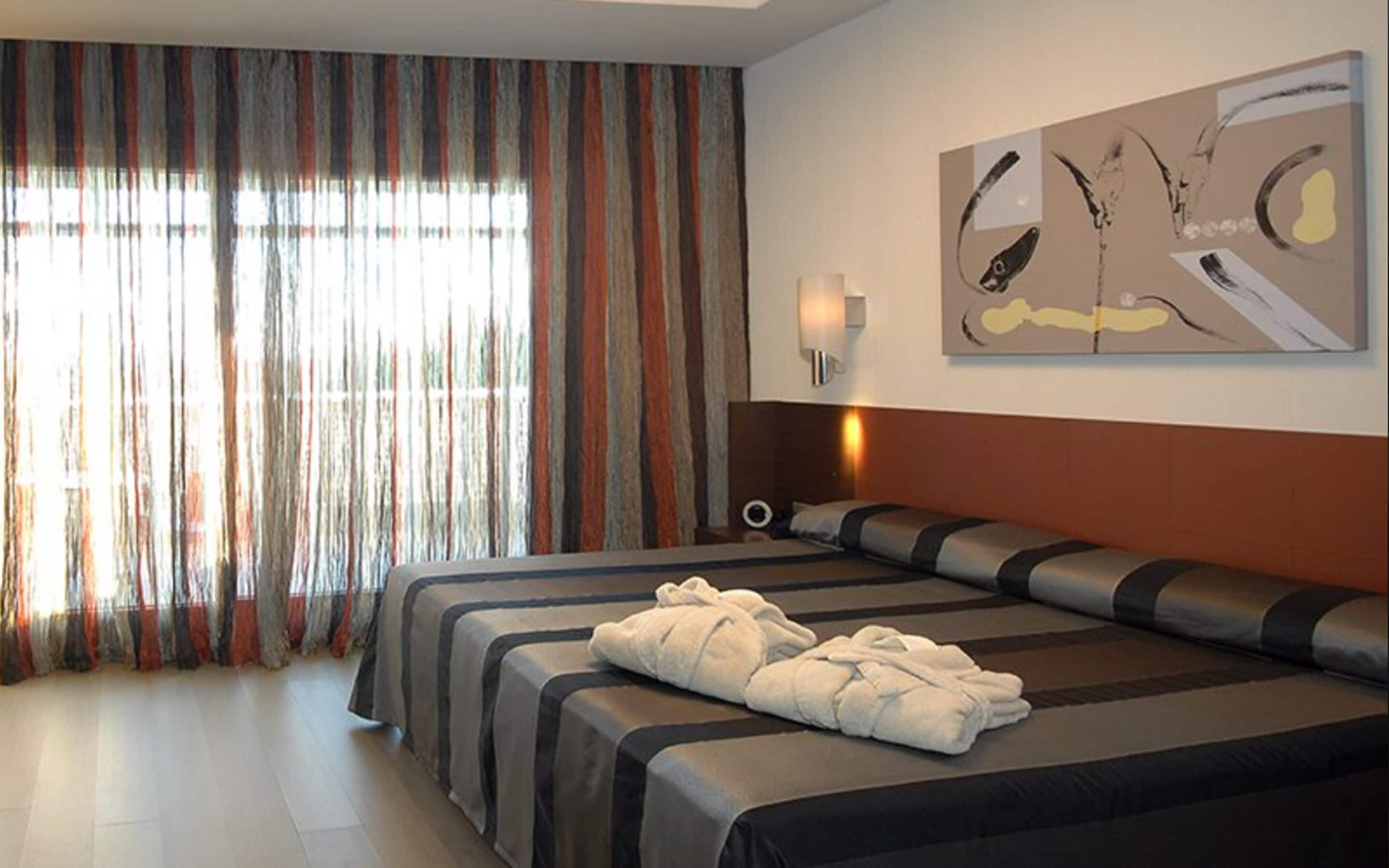 Una camera d'albergo con due letti, una grande finestra con tende, una lampada a parete e un quadro.