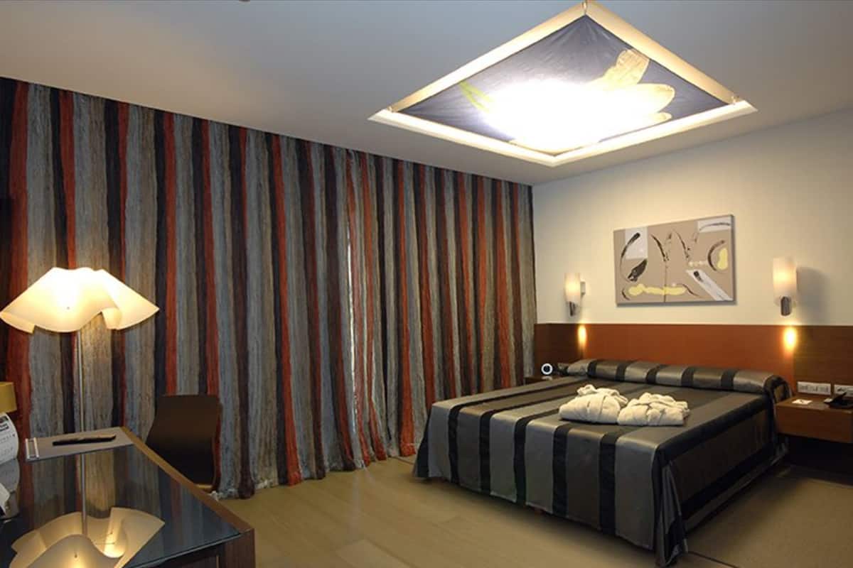 A hotel room with a bed, a desk, a lamp, a ceiling light, curtains, and a wall art piece.