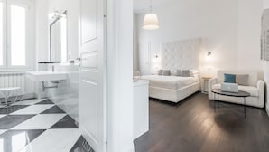 A modern hotel room with a checkered floor, a white bed, a sofa, a small table with a laptop, and a bathroom with a white sink and mirror.
