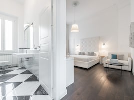 Habitación de hotel moderna con suelo a cuadros, cama blanca, sofá, una pequeña mesa con un ordenador portátil y baño con lavabo y espejo blancos.